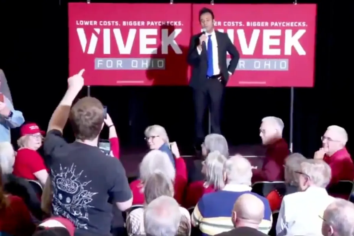 Man speaking on stage before seated audience at campaign event with red Ohio campaign backdrop.