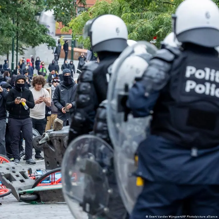 Riot police in helmets and shields face a crowd of masked and hooded protesters on a street.