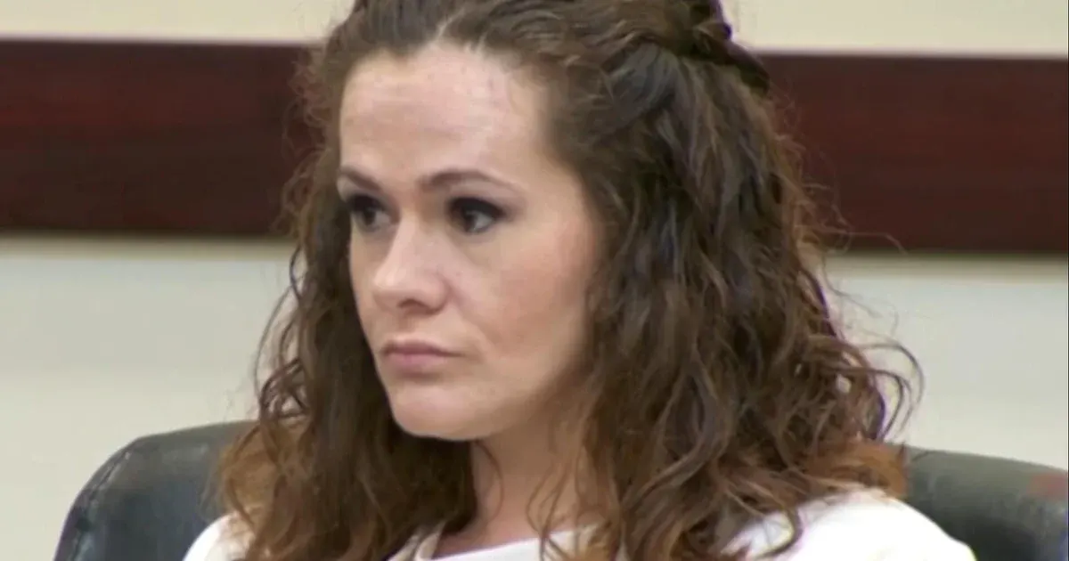 Woman with wavy brown hair sits in a courtroom-style setting, looking off to the side.