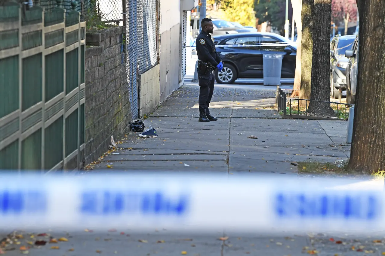 Police officer in blue gloves stands on a sidewalk behind crime scene tape, debris visible.