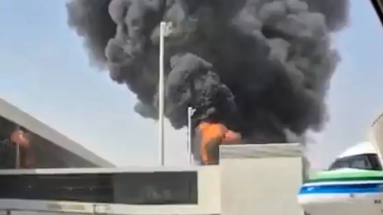 Large black smoke plume rising behind airport structures and an aircraft, with orange flames visible.