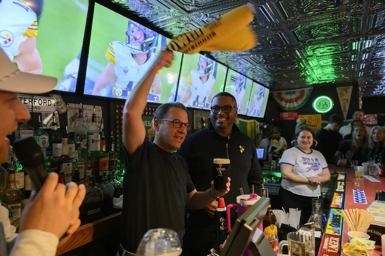Two smiling men at a bar, one waving a yellow towel, with football game on screens behind.