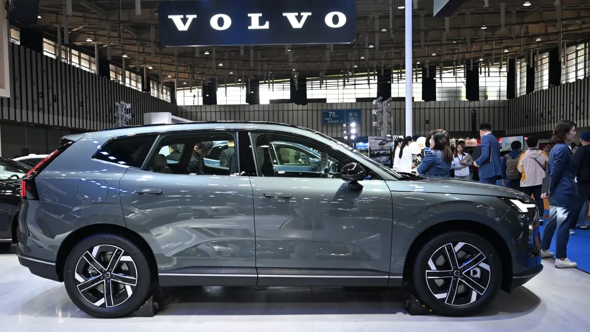 Grey Volvo SUV displayed at an auto show, Volvo logo sign overhead, visitors behind.