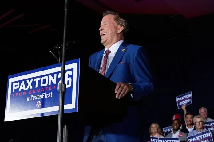 Smiling man in blue suit speaks at podium with Paxton for Senate campaign signs behind him.