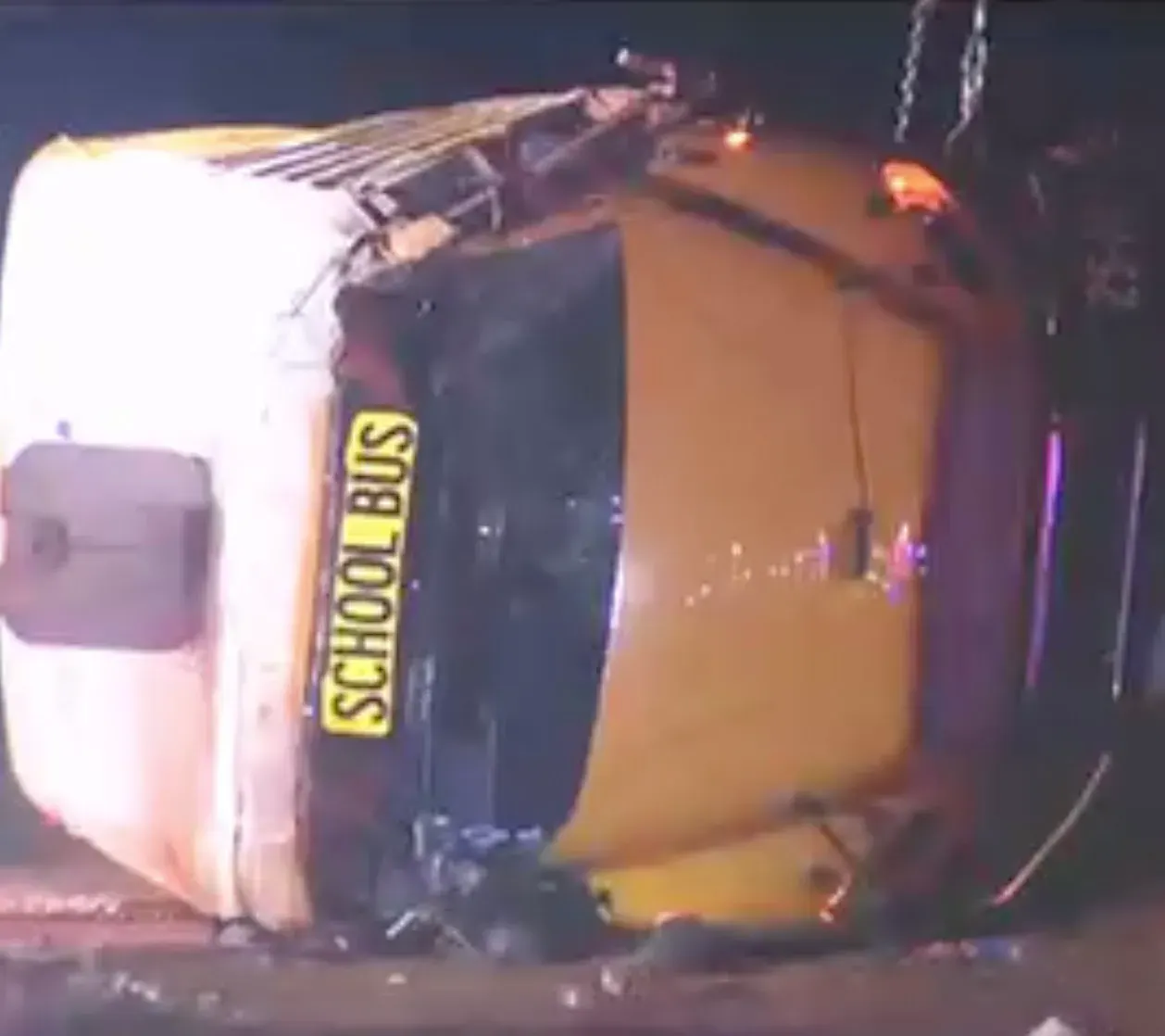 Yellow school bus lying on its side at night, damaged, with debris on the roadway.