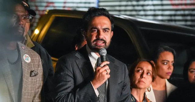 Man speaking into a microphone at a nighttime outdoor event, flanked by people.