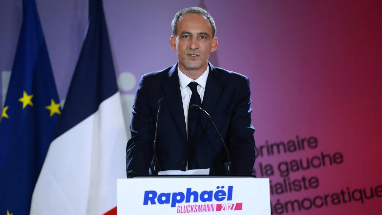 A man speaks at a podium bearing campaign branding, with French and EU flags behind him.