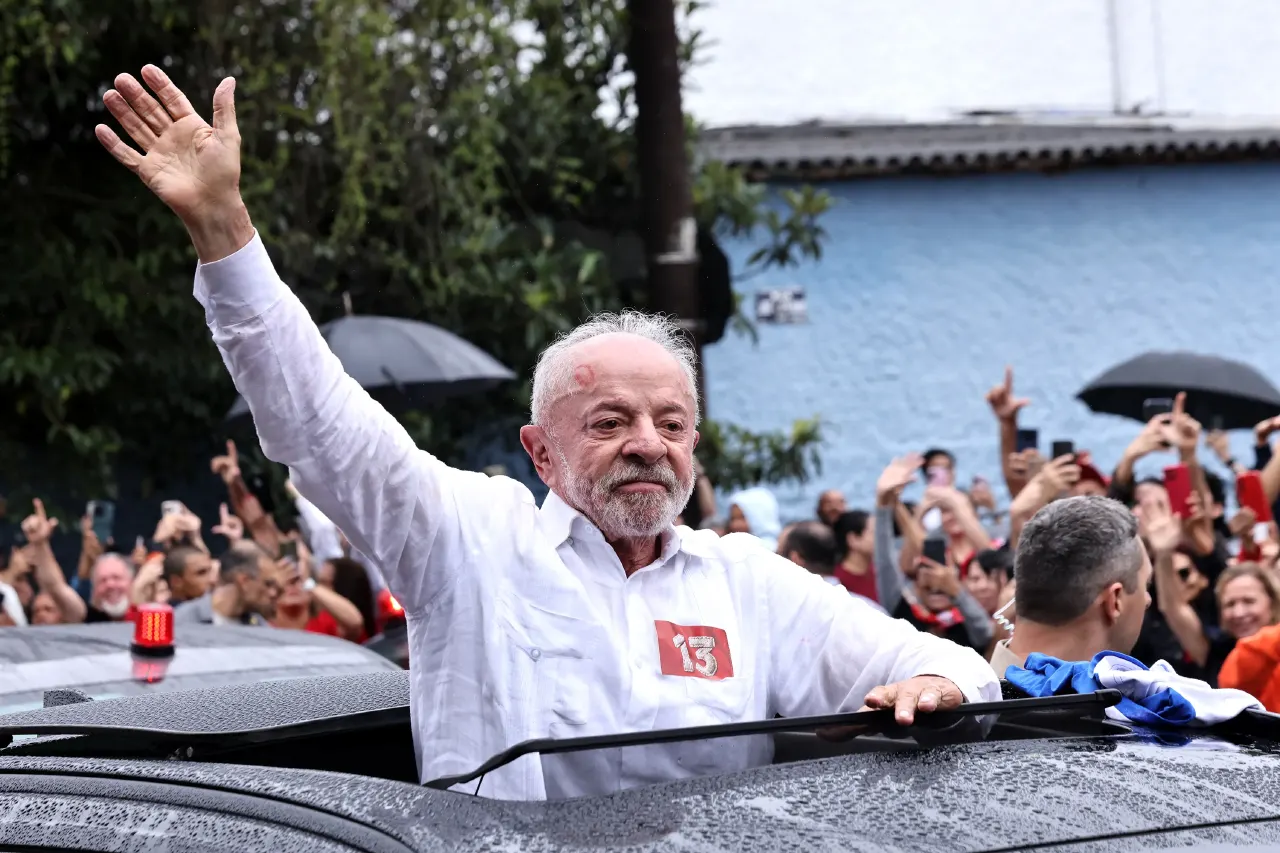 White-haired bearded man in white shirt with number 13 waves from open car to crowd.