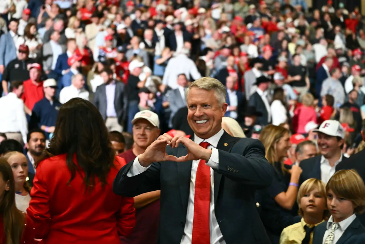 Smiling man in suit and red tie making heart gesture at packed rally crowd wearing red caps