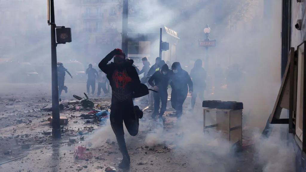Masked figures running through tear gas smoke amid debris on a Paris street.