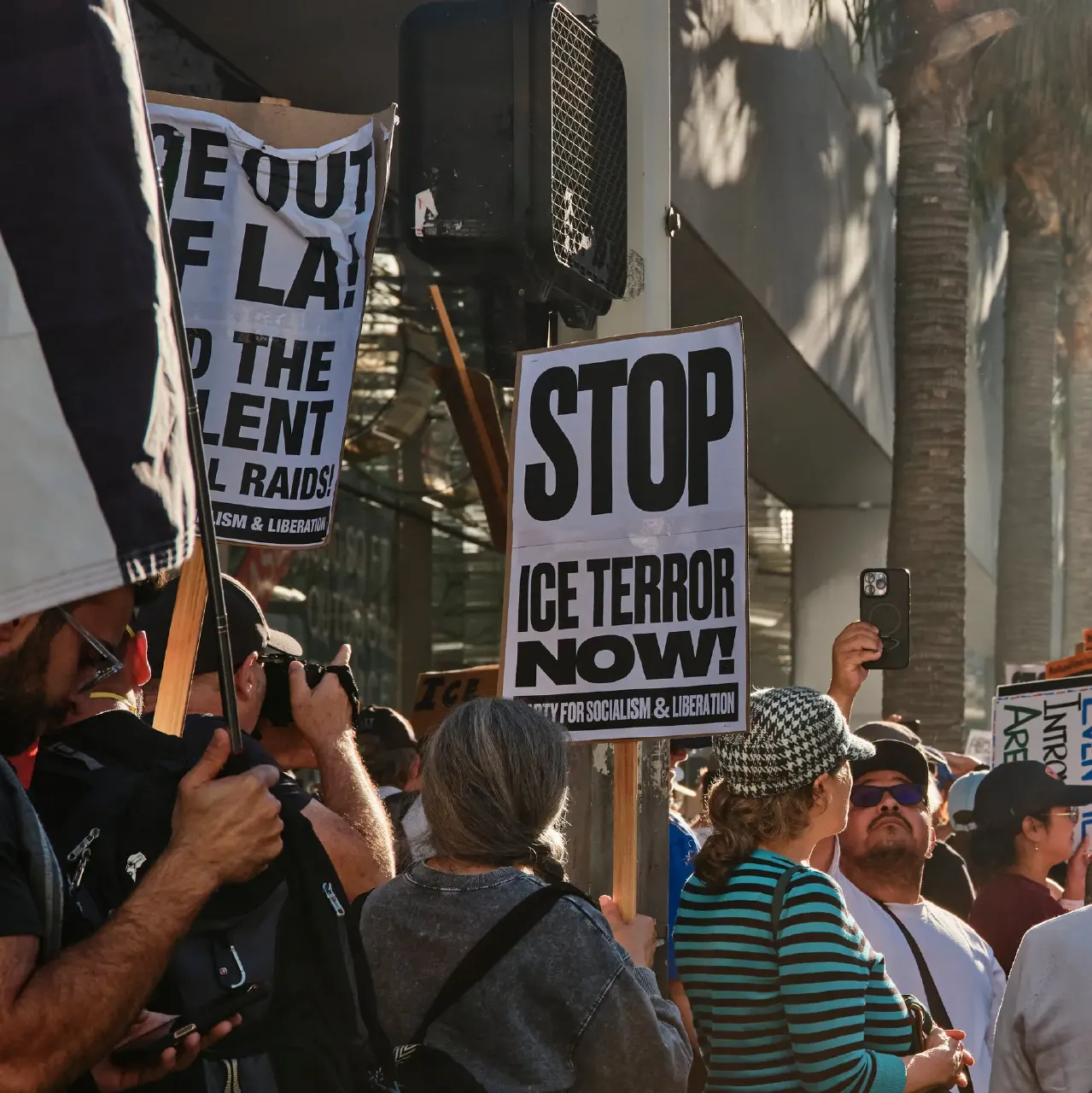 Protesters with signs reading 'STOP ICE TERROR NOW' and 'Raids' at a street demonstration.