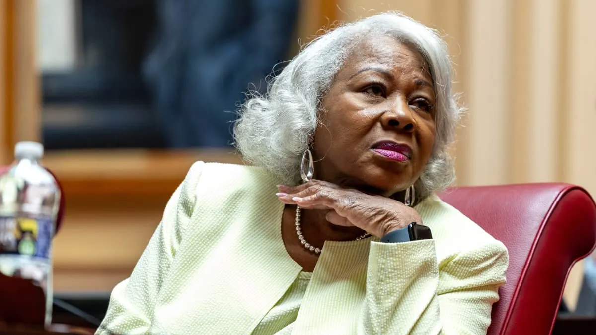 Older woman with white hair in pale green jacket, resting chin on hand in a legislative chamber.