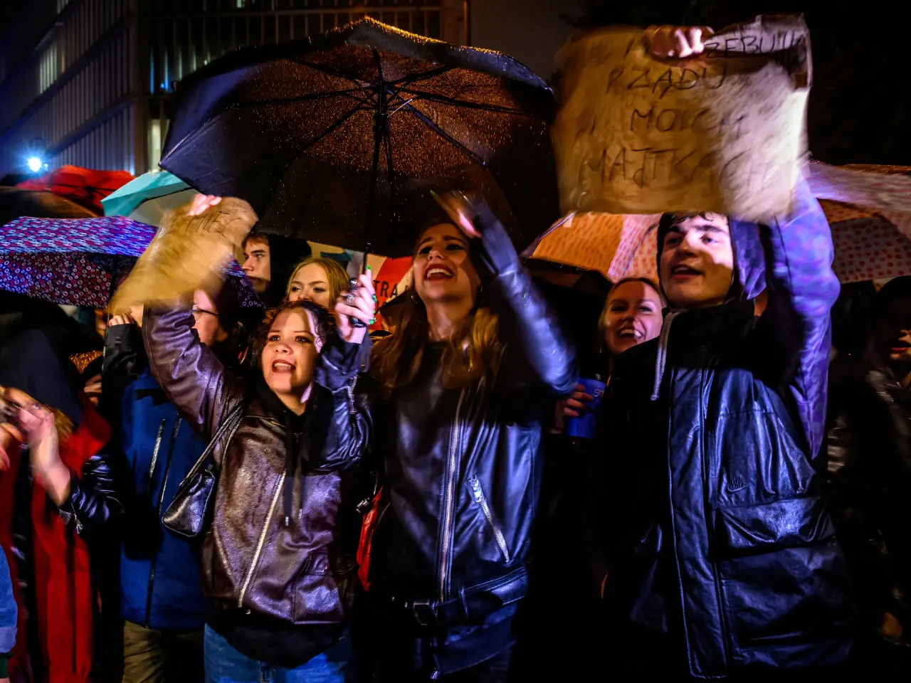 Crowd of people under umbrellas at night, shouting, one holding a handwritten protest sign.