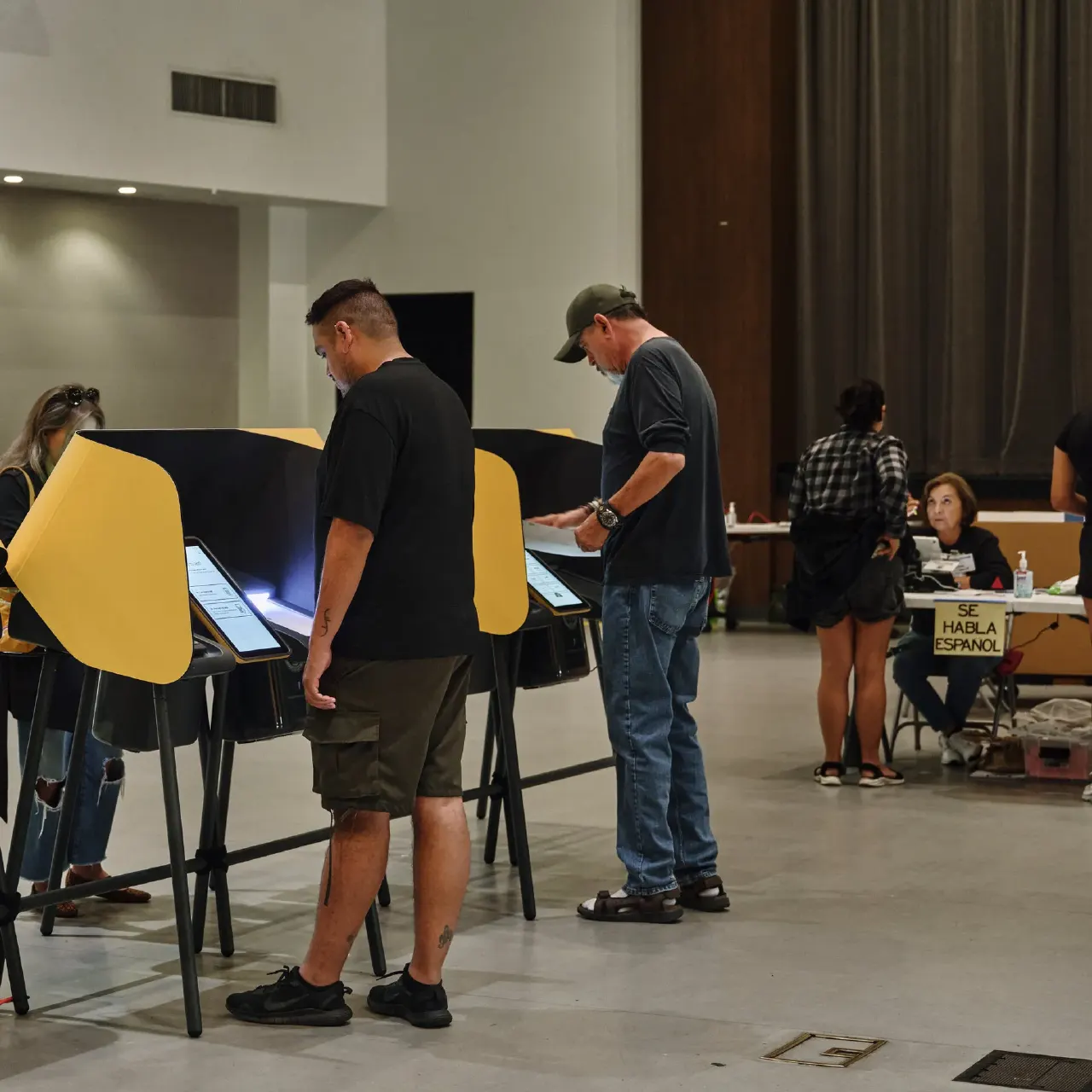 Voters use privacy booths at a polling place, with a Spanish-language sign and poll worker.