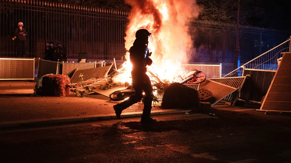 Person in dark gear walks past a large fire burning debris and barricades at night on a street.