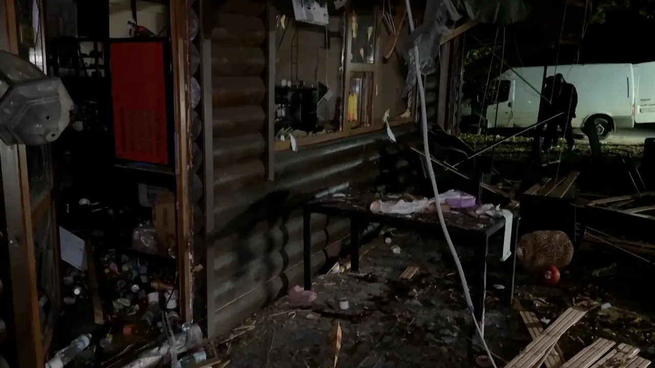 Night view of a damaged, debris-strewn wooden structure with broken glass and a white van in background.