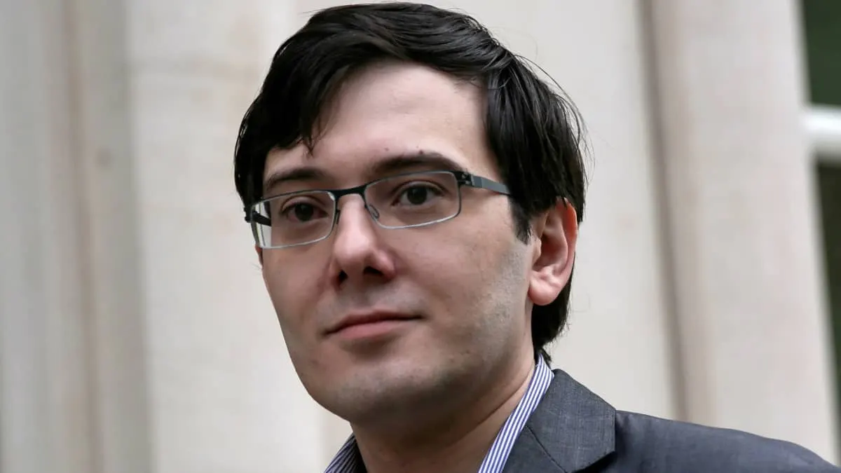 Man in glasses and grey blazer in front of pale stone columns, looking at camera.
