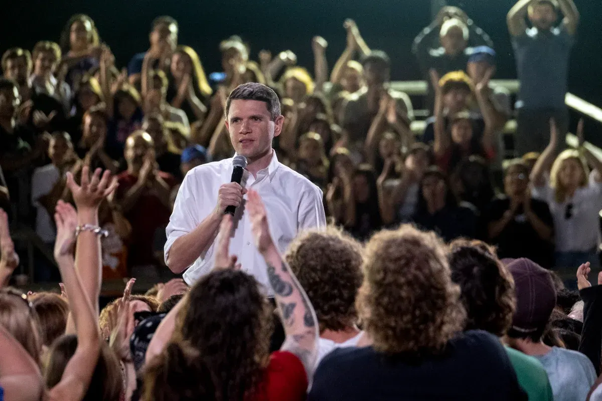 A man in a white shirt speaks into a microphone to a cheering crowd with raised hands.