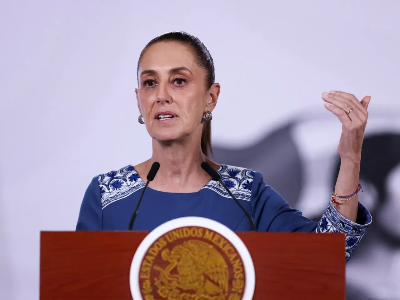 A woman in a blue embroidered blouse speaks at a podium bearing the Mexican national seal.
