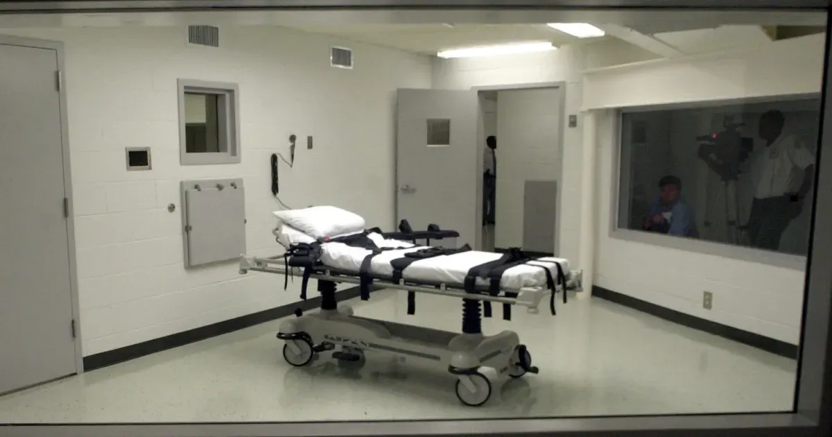 Execution chamber with gurney and restraints, viewed through glass, with observers behind a window.