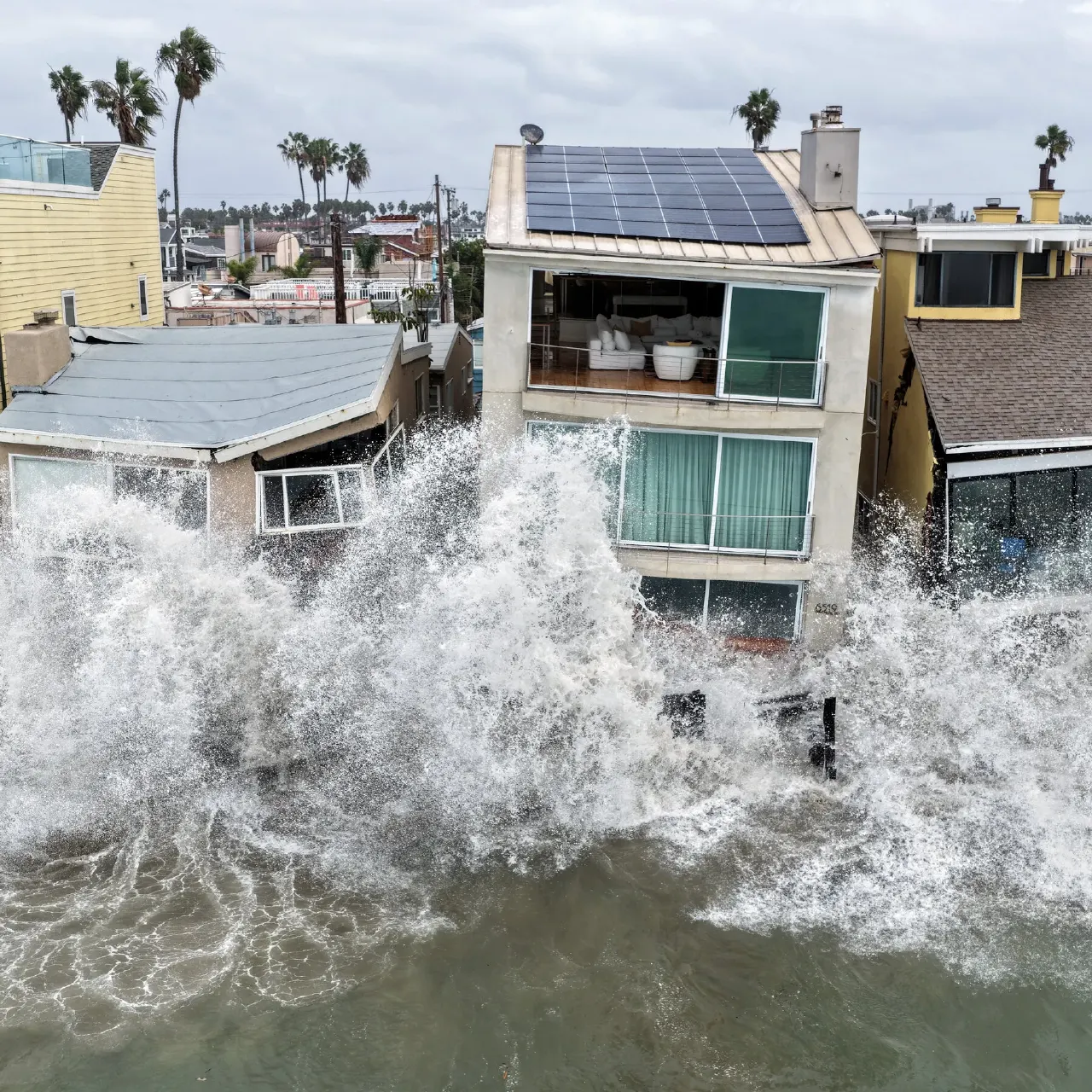 Large waves crash over beachfront homes in an aerial view of coastal flooding.