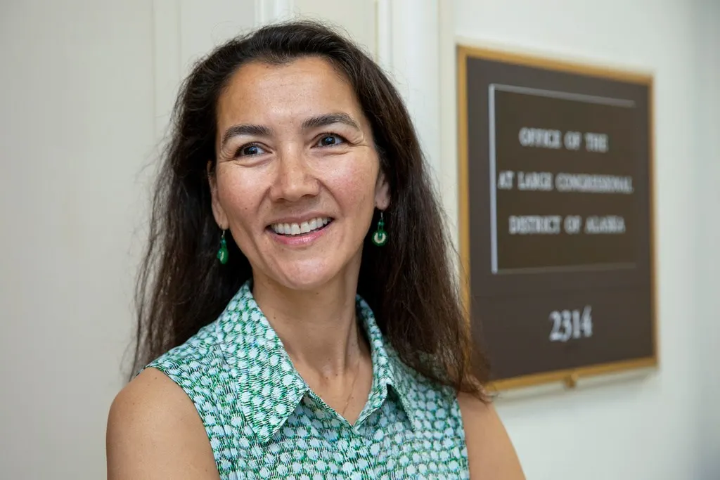 Smiling woman with long dark hair beside an office sign reading Congressional District of Alaska.