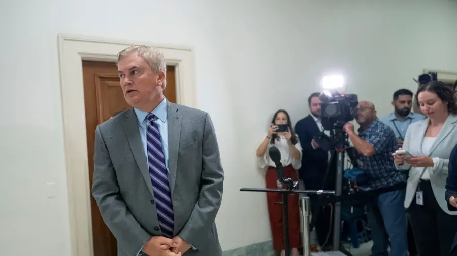 Man in grey suit speaks at a microphone while reporters and cameras film in a hallway.