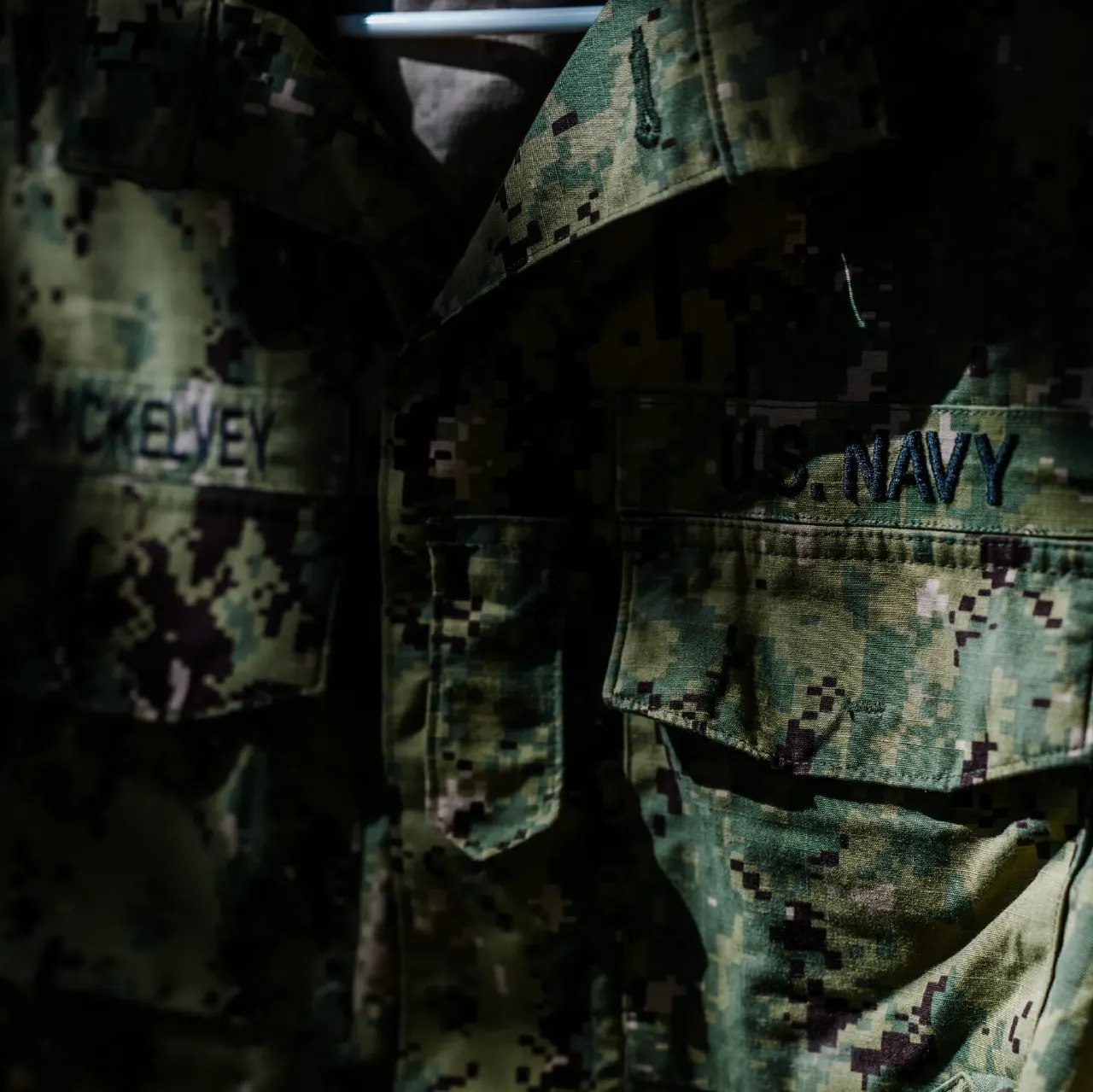 Dim close-up of camouflage uniforms with U.S. Navy and a partial name tape.