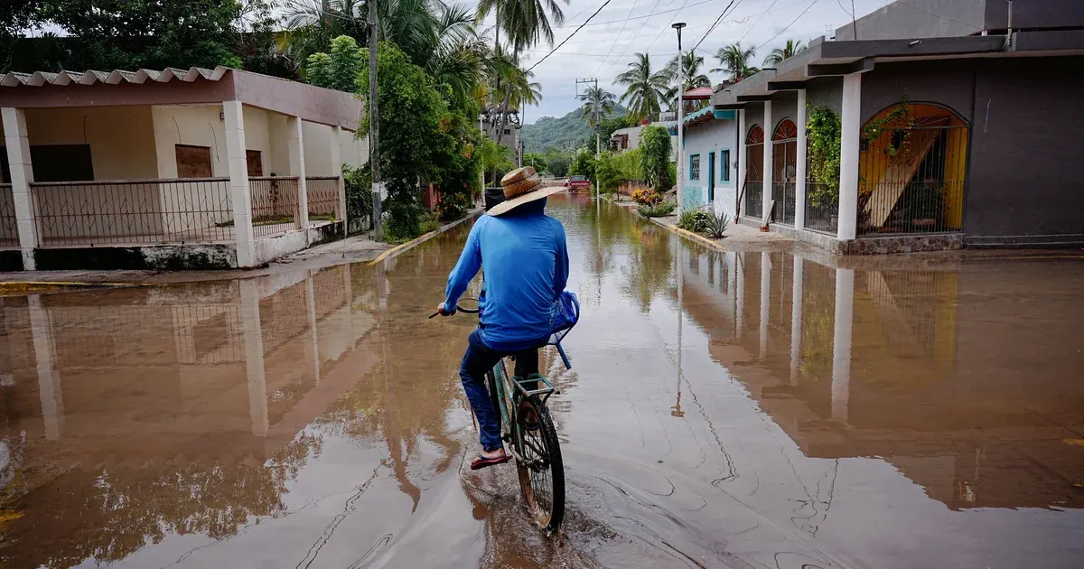 Man in straw hat cycles through flooded street past houses after storm, water reaching the wheels.