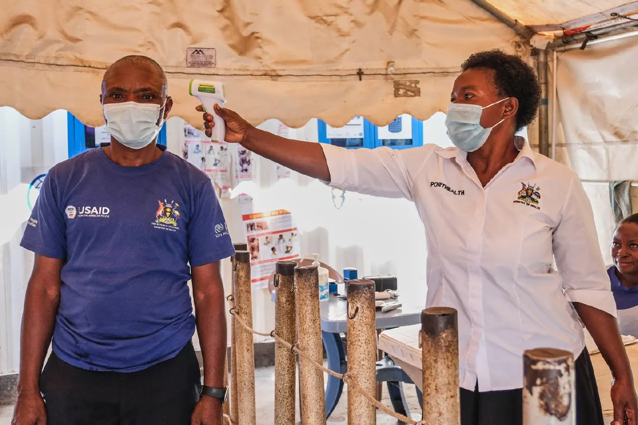 Health worker in face mask uses infrared thermometer on a man at a screening tent with USAID logo