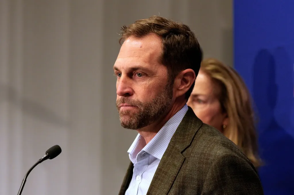 Bearded man in tweed jacket speaking at a microphone, a woman behind him