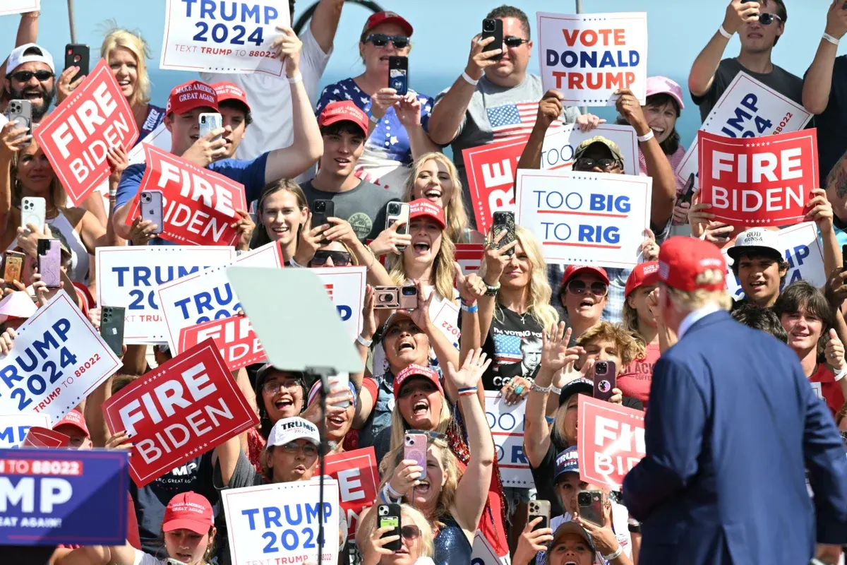 Crowd of supporters holding Trump 2024 and Fire Biden signs and Vote Donald Trump placards, many raising phones, with a man in a suit and red cap seen from behind.