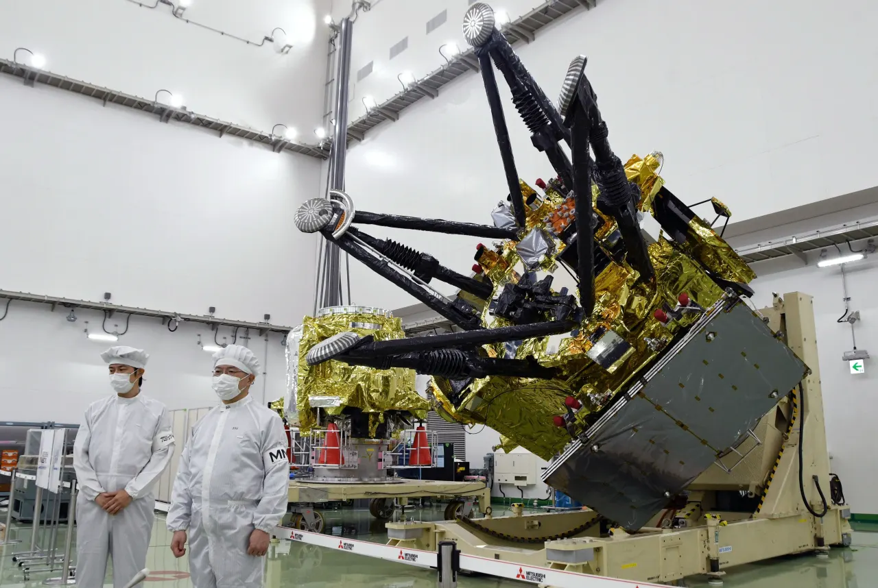 Two technicians in cleanroom suits watch a gold-foiled spacecraft with folded black arms being prepared.