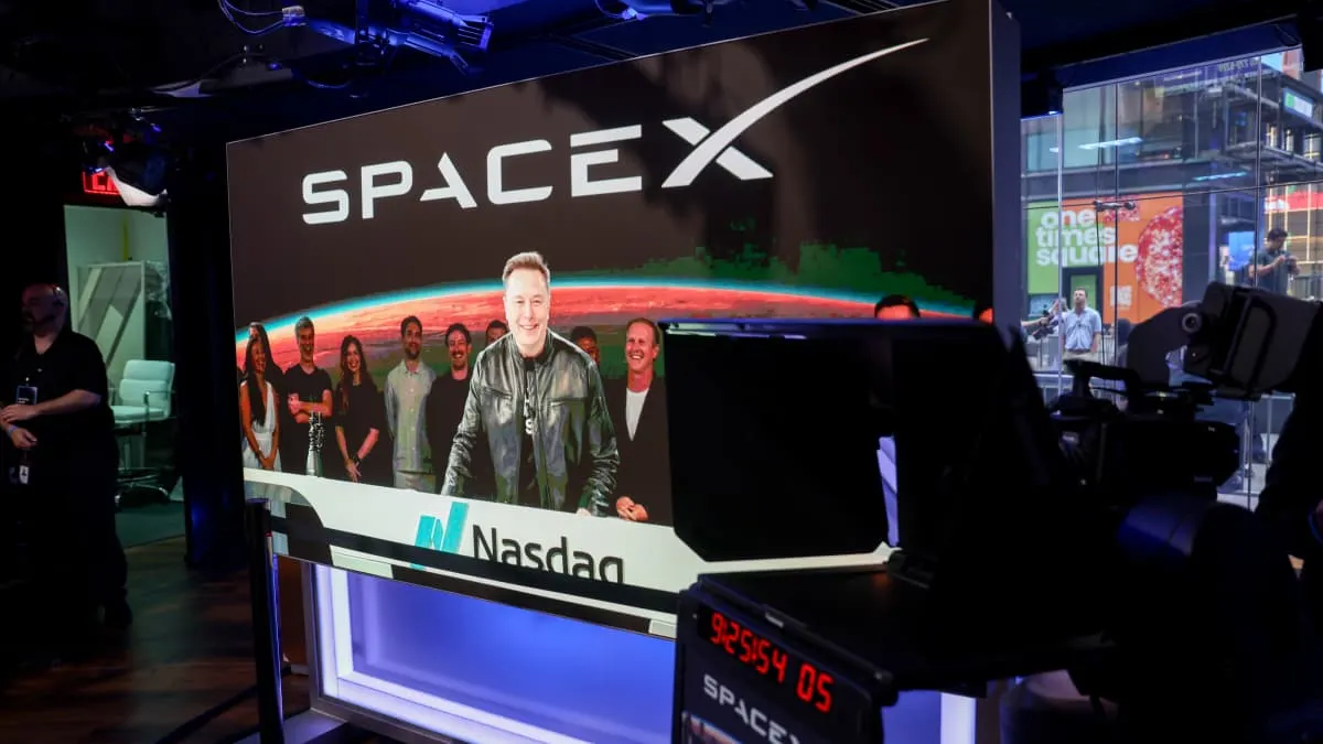 Man in leather jacket smiling on screen behind SpaceX logo, Nasdaq branding, camera equipment in foreground.
