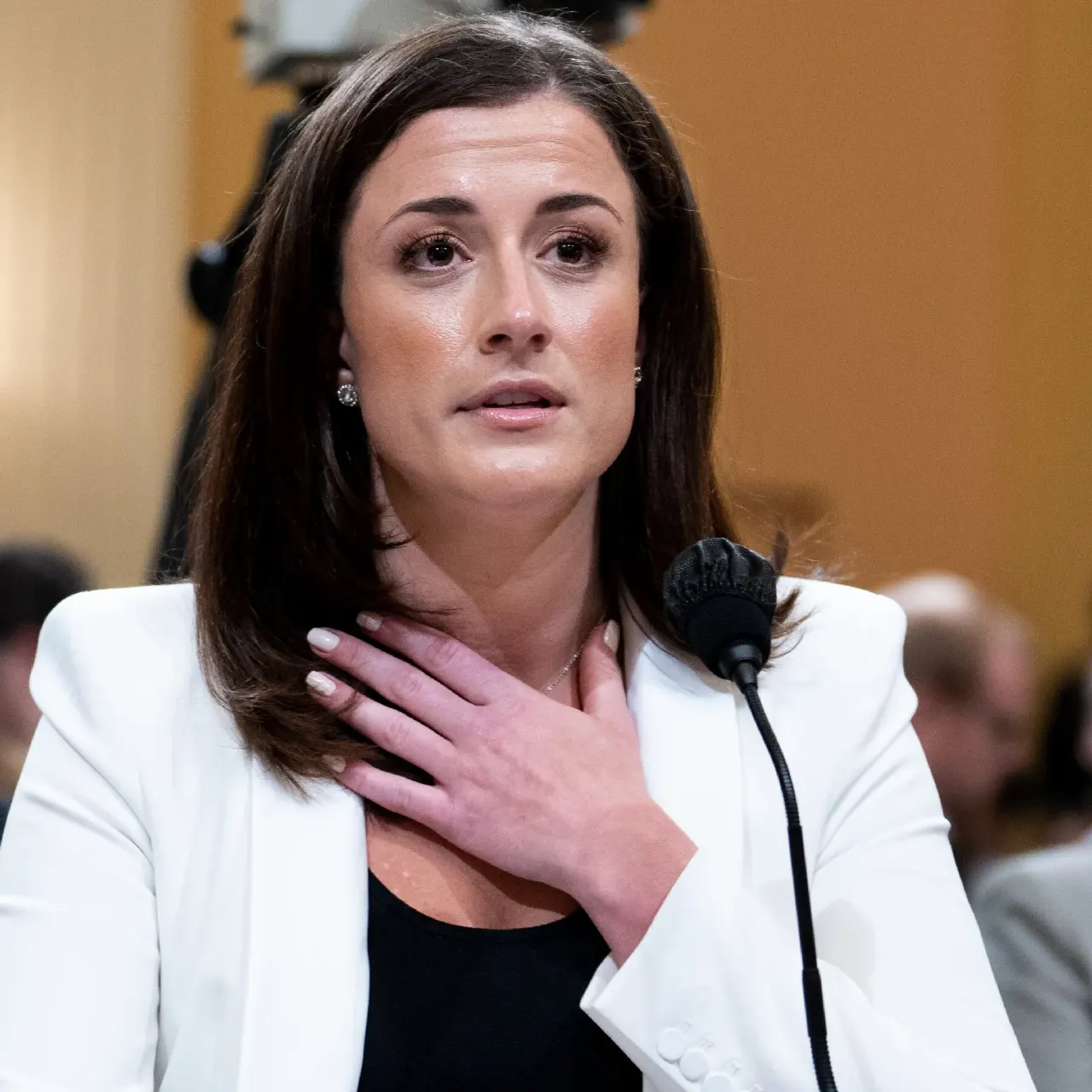 Woman in white blazer, hand on chest, speaking into a microphone at a hearing.