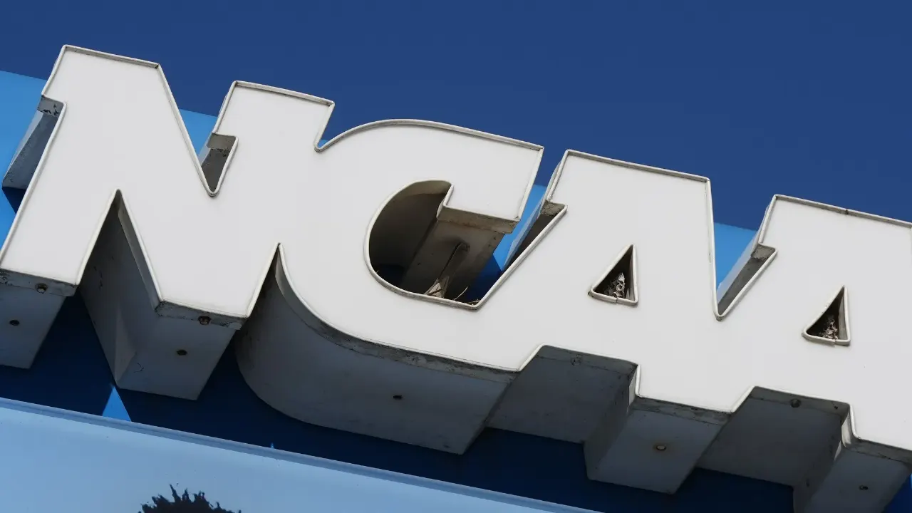 Large white NCAA letters mounted on a building against a blue sky, shot from below.