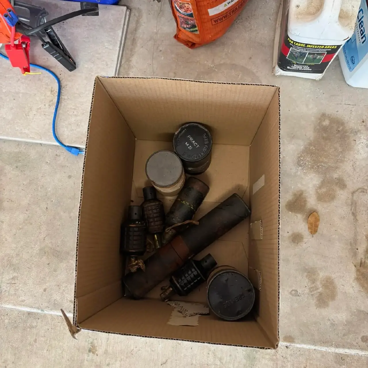 Cardboard box on concrete holding several old military grenades and canisters, overhead view.