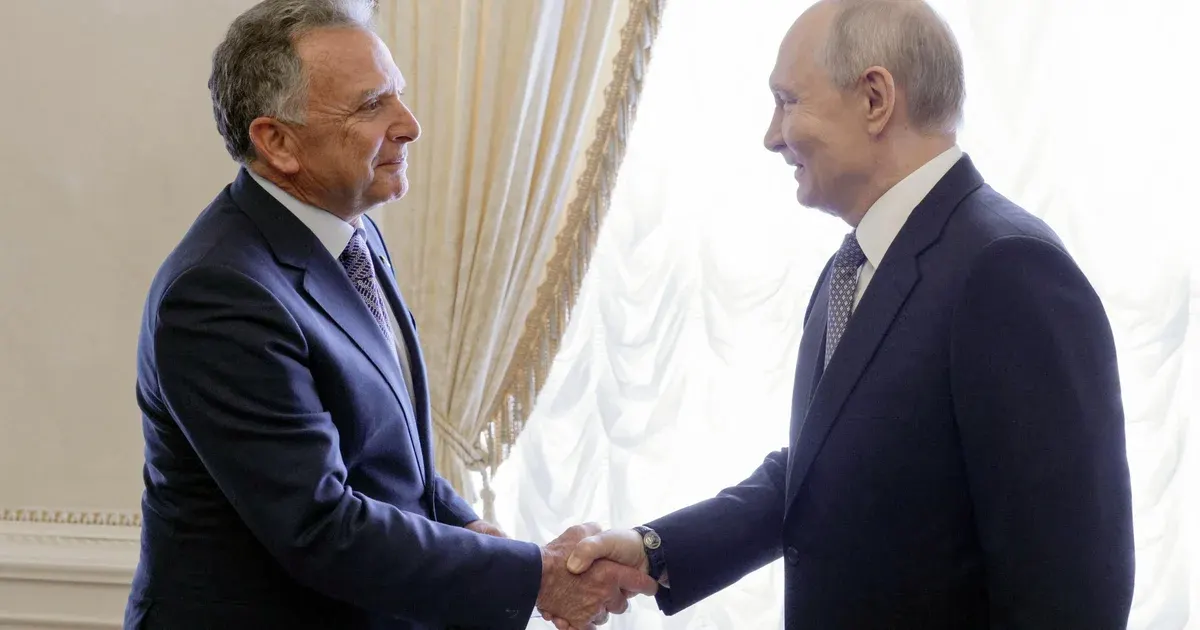 Two men in dark suits shake hands in a formal room with white drapes.