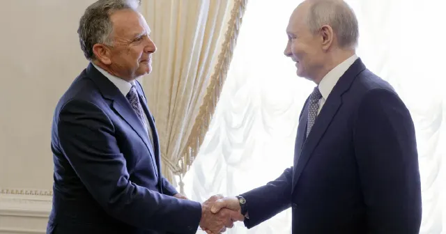 Two men in dark suits shake hands in a formal room with white drapes.