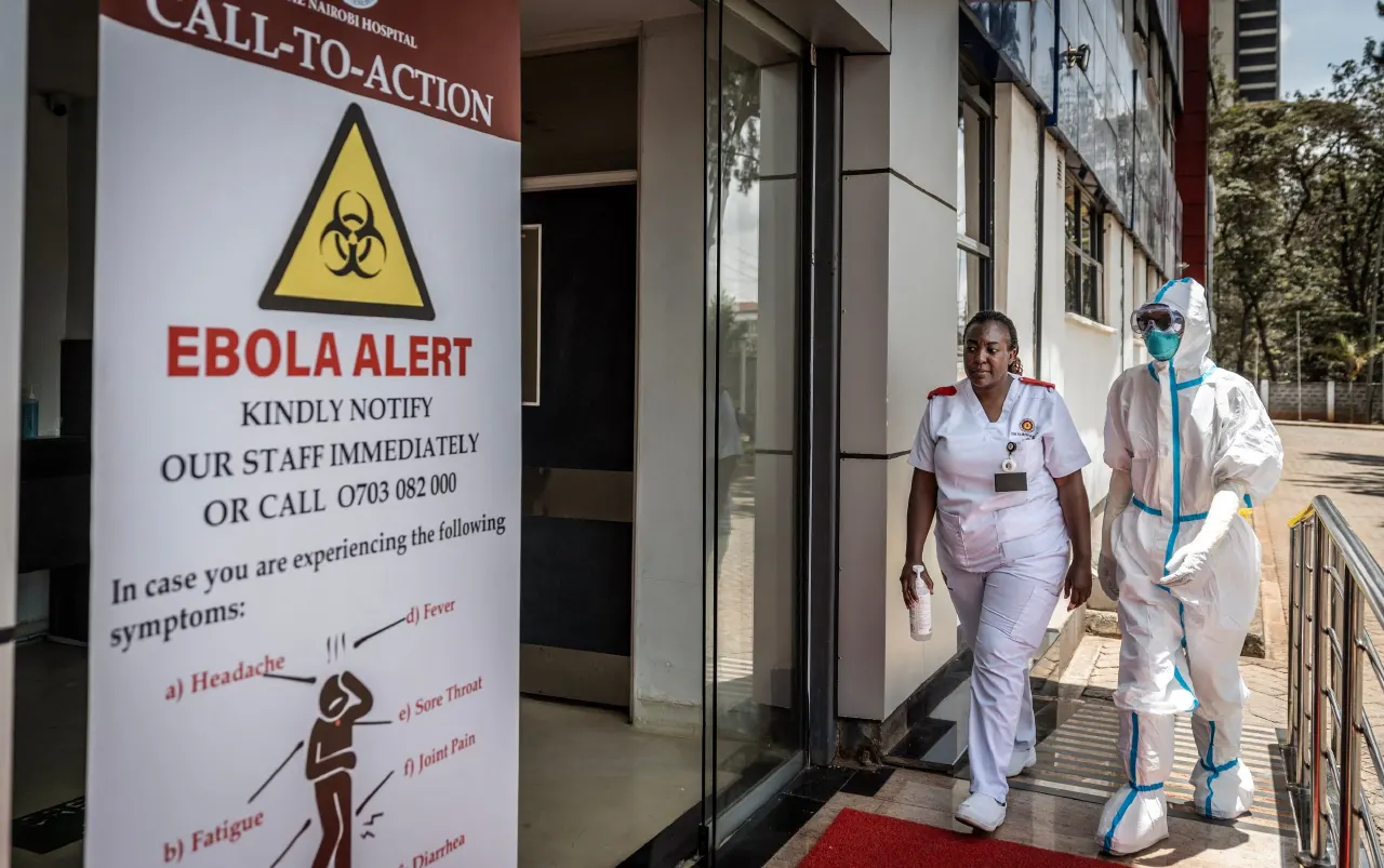 Hospital entrance with Ebola alert sign; nurse and worker in full protective suit walking out.