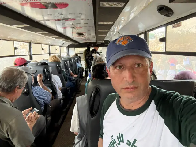 Man in cap and green-and-white shirt taking selfie on crowded bus, armed soldier visible behind.