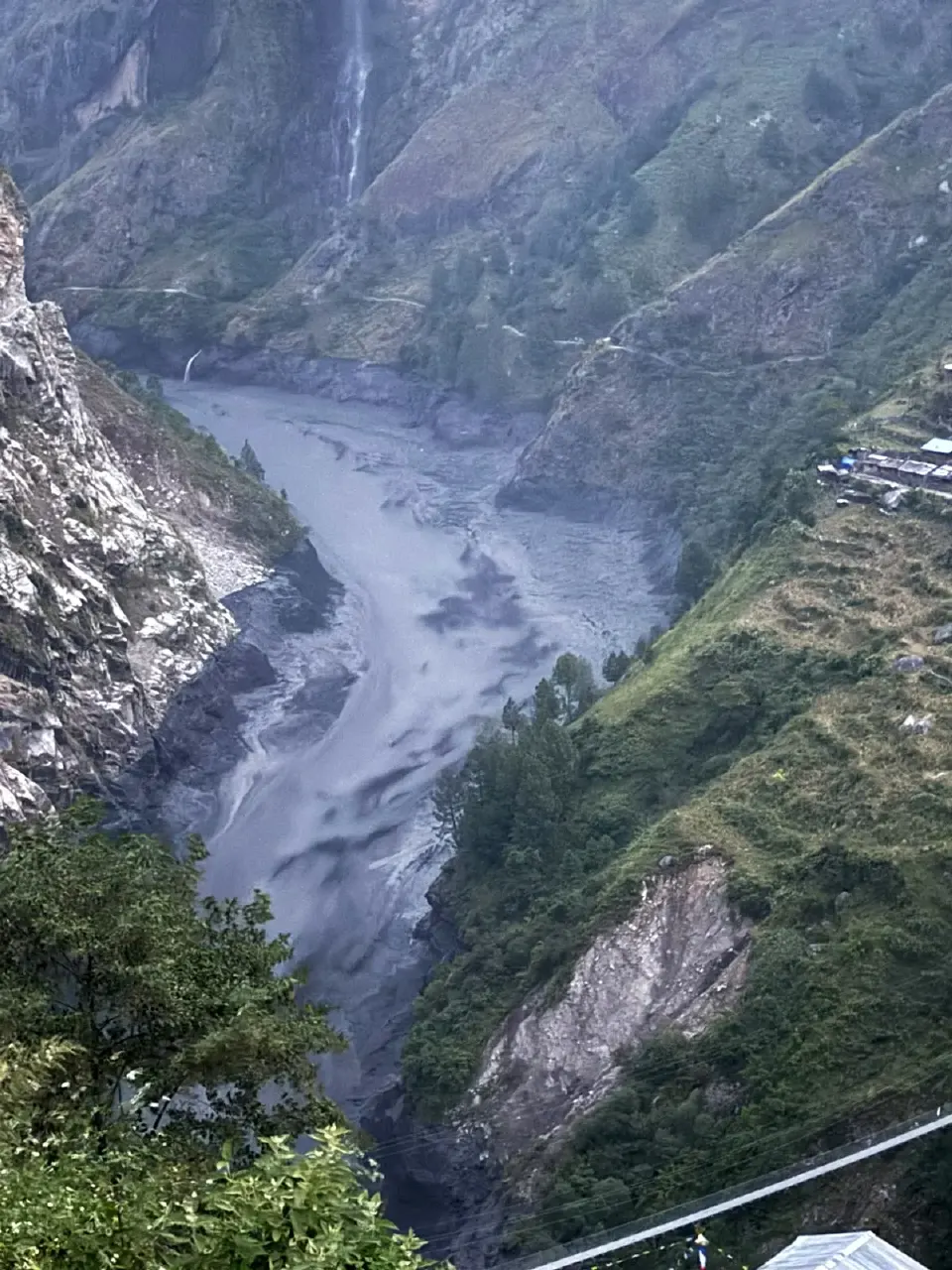 Muddy grey floodwater rushing through a steep mountain valley, with buildings on the hillside at right.