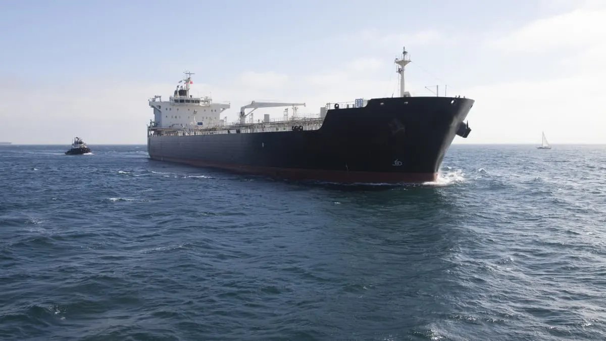 Large black oil tanker at sea with a tugboat and a distant sailboat on calm water.