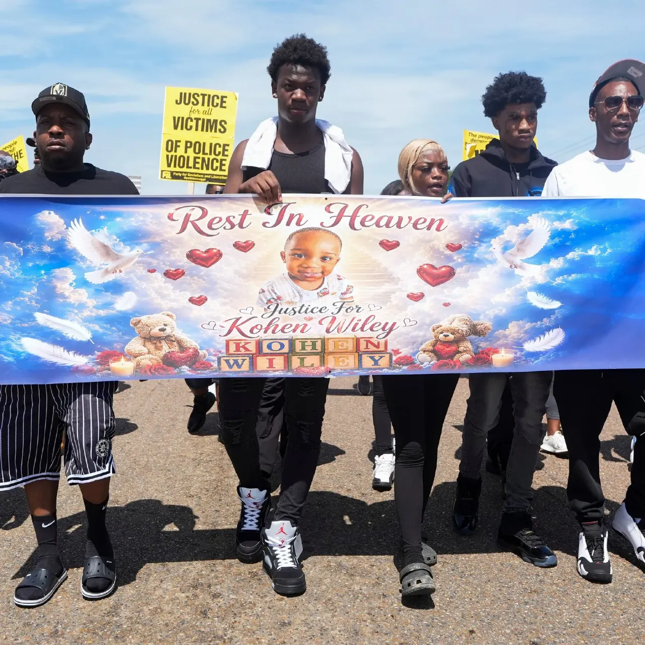 Marchers hold a 'Rest In Heaven' memorial banner for a young child, with protest signs behind.