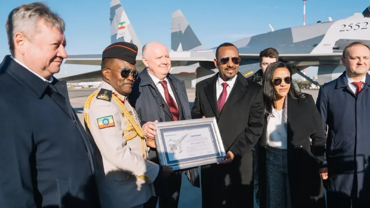 Officials in formal dress presenting a framed certificate before a Su-35 jet on an airfield tarmac.