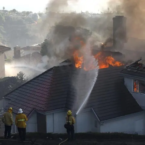 Firefighters in yellow gear spray water on a burning house roof with thick smoke rising.