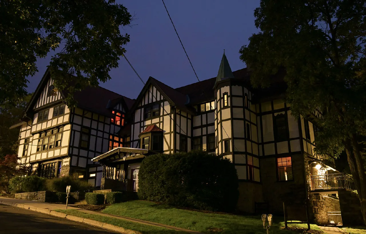 Tudor-style fraternity house with timber framing illuminated at dusk, surrounded by trees and shrubs.