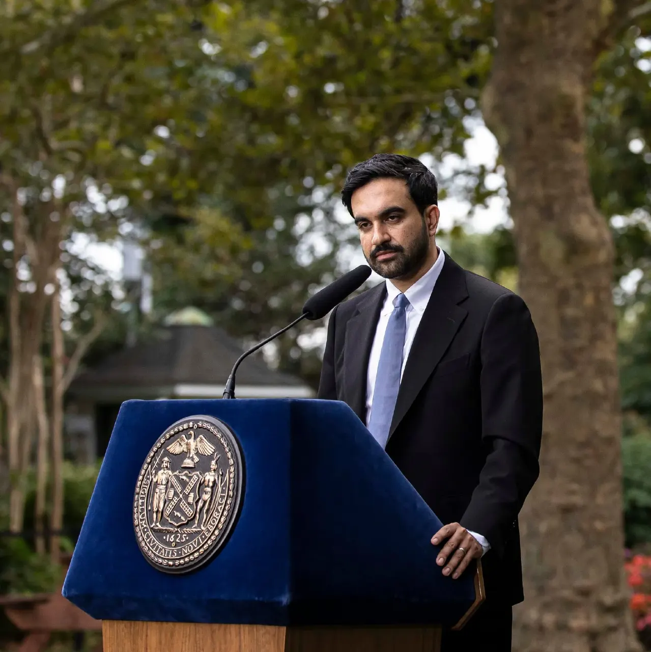 A man in a dark suit speaks at a blue podium bearing a city seal, outdoors among trees.