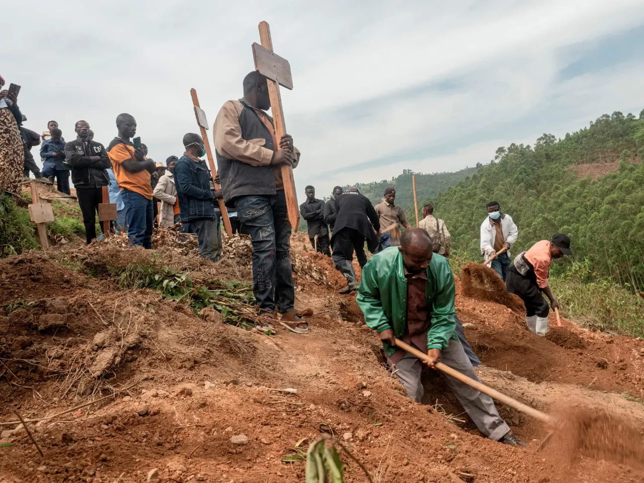Men digging graves on a hillside, one holding a wooden cross, others watching, masks visible.