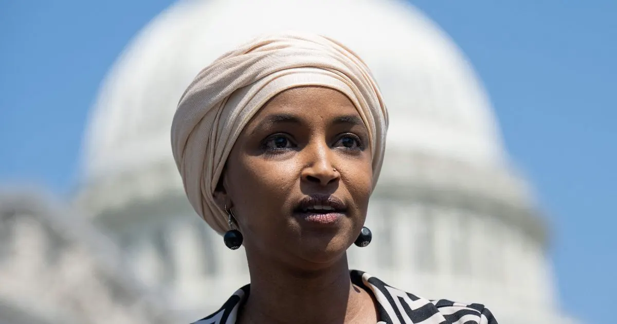 Woman in a cream head wrap and patterned top speaking, with a blurred white domed building behind her.